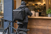 Synthetic editorial illustration. Anonymous bicycle courier seen entirely from behind at a restaurant meal pickup counter, with plain insulated bag and sealed takeaway food containers; no named delivery service.