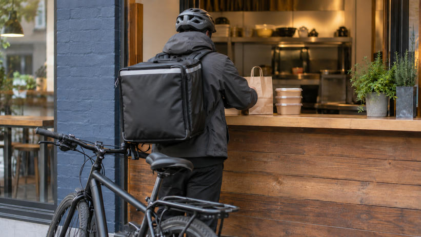 Synthetic editorial illustration. Anonymous bicycle courier seen entirely from behind at a restaurant meal pickup counter, with plain insulated bag and sealed takeaway food containers; no named delivery service.