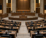 Synthetic editorial illustration. A few attendees viewed from behind sit among semicircular rows of wooden desks in a large assembly hall. The dais is empty, and all desk and wall displays are dark under warm institutional light.