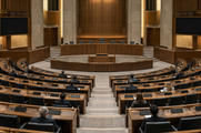 Synthetic editorial illustration. A few attendees viewed from behind sit among semicircular rows of wooden desks in a large assembly hall. The dais is empty, and all desk and wall displays are dark under warm institutional light.