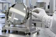 Synthetic editorial illustration. A white-gloved hand adjusts a plain metal mount holding a large circular mirror. The precision module sits on a perforated cleanroom table and reflects soft overhead light without faces.