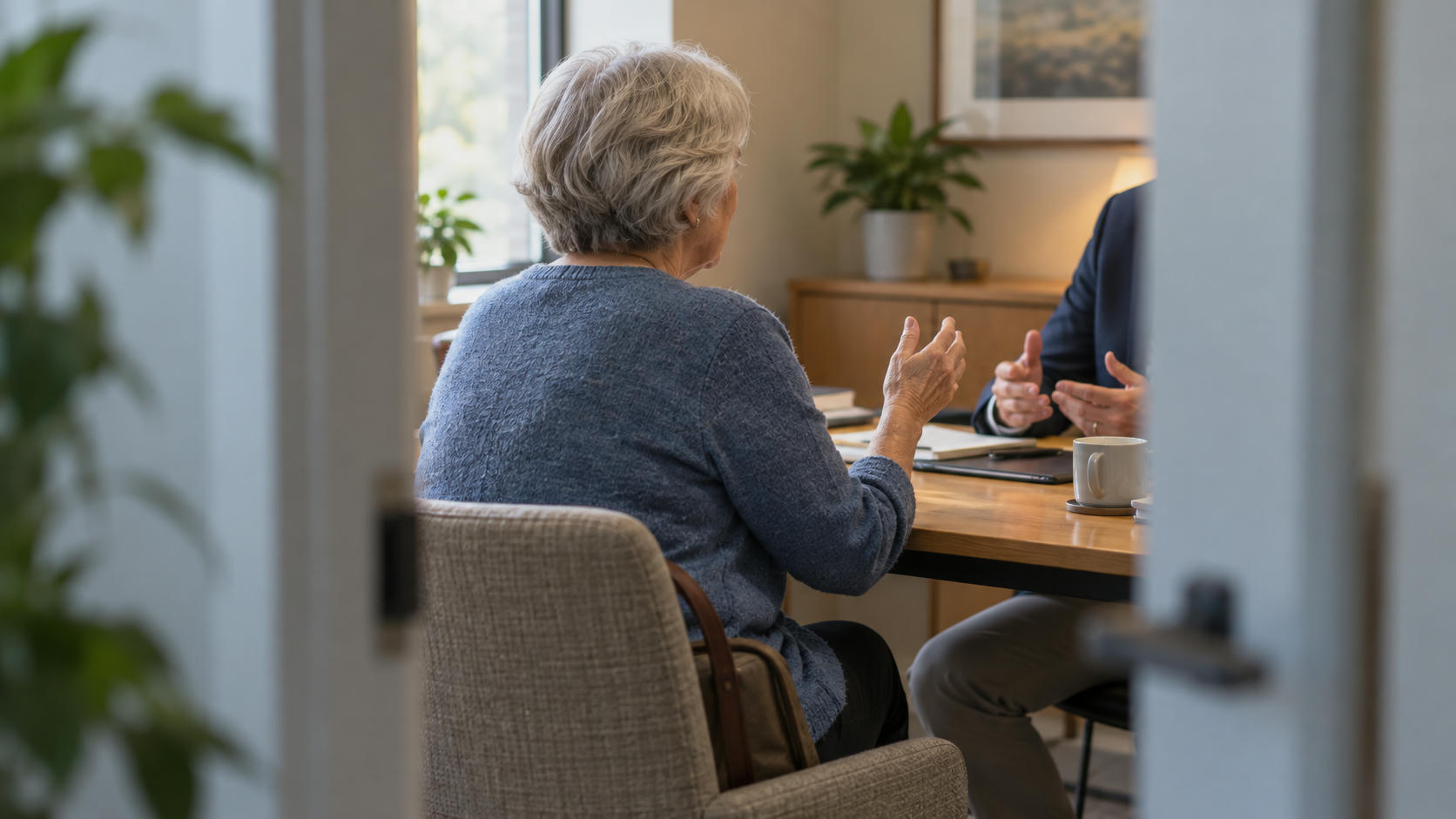 Synthetic editorial illustration. A grey-haired older adult viewed from behind at a consultation table, engaged in conversation and natural hand gestures with a secondary adviser whose head is outside the frame. Understated documents remain secondary; no open blank book or reading pose.