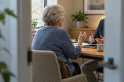 Synthetic editorial illustration. A grey-haired older adult viewed from behind at a consultation table, engaged in conversation and natural hand gestures with a secondary adviser whose head is outside the frame. Understated documents remain secondary; no open blank book or reading pose.