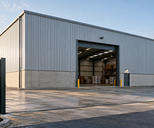 Synthetic editorial illustration. One anonymous modern warehouse facade and open loading portal with modest unmarked freight inside, naturally framed by entrance fencing.