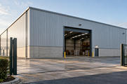 Synthetic editorial illustration. One anonymous modern warehouse facade and open loading portal with modest unmarked freight inside, naturally framed by entrance fencing.