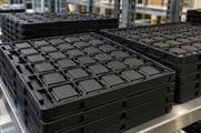 Synthetic editorial illustration. Stacks of black molded trays hold orderly rows of plain dark chip packages with fine metal contacts. The trays sit on a stainless factory shelf in soft industrial light.