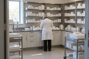 Synthetic editorial illustration. A hospital pharmacy room. A pharmacist in a white coat seen from behind prepares trays at a counter, surrounded by shelves and trolleys of plain white boxes and bottles.