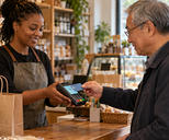 Synthetic editorial illustration. A shopper presents a plain payment card to a handheld terminal held by a shopkeeper.