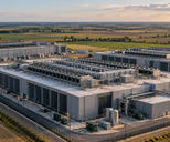 Synthetic editorial illustration. A data-centre campus sits among flat farmland. A low aerial view shows large windowless buildings, rooftop chillers, pipework and fenced service roads.