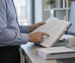 Synthetic editorial illustration. A cropped adult reviews an open populated report beside a powered portrait monitor, indicating a paragraph by hand.
