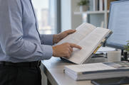 Synthetic editorial illustration. A cropped adult reviews an open populated report beside a powered portrait monitor, indicating a paragraph by hand.