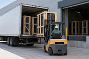 Synthetic editorial illustration. An industrial loading dock. A forklift operator seen from behind handles a wooden-crated server rack beside an open truck, with more crated racks inside the truck and building.