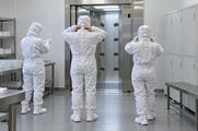 Synthetic editorial illustration. Three workers viewed from behind adjust white cleanroom suits in a plain gowning room. Blank lockers and metal benches flank a stainless air-shower doorway with circular nozzles.