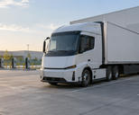 Synthetic editorial illustration. Generic unbranded electric cargo tractor and plain trailer at an anonymous freight depot beside ordinary charging infrastructure.