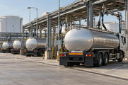 Synthetic editorial illustration. A fuel-loading depot. Silver tanker trucks stand beneath a canopy of loading arms and pipework, with large storage tanks beyond and plain black mudflaps above the concrete yard.