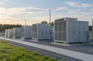 Synthetic editorial illustration. A generic row of plain battery-storage enclosures and utility connections in a grid-scale energy yard; not a named product or facility.