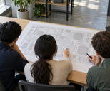 Synthetic editorial illustration. Three people viewed from above and behind examine a large paper circuit pattern of rectangular components and fine connecting traces. Two plain closed laptops sit on the wooden table in daylight.