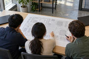 Synthetic editorial illustration. Three people viewed from above and behind examine a large paper circuit pattern of rectangular components and fine connecting traces. Two plain closed laptops sit on the wooden table in daylight.