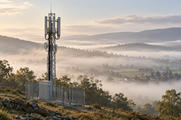 Synthetic editorial illustration. A cellular relay mast and its base cabinet stand above a misty rural valley.