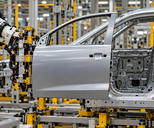 Synthetic editorial illustration. A robot supports a silver front door with a left-sloping front window edge and right-hand handle beside the corresponding near-side front body opening.