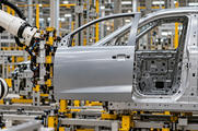 Synthetic editorial illustration. A robot supports a silver front door with a left-sloping front window edge and right-hand handle beside the corresponding near-side front body opening.