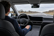 Synthetic editorial illustration. Viewed from the back seat, a driver rests both hands on their legs away from a plain steering wheel. Dashboard displays are dark, and a straight empty motorway extends through the windscreen under grey daylight.