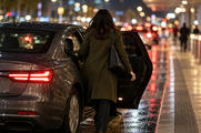 Synthetic editorial illustration. A city pickup curb. A rear-facing passenger in an olive coat steps through a sedan's open rear door holding a dark phone, as blurred traffic lights reflect across the wet pavement at night.
