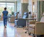 Synthetic editorial illustration. A nurse seen from behind walks through a bright treatment room lined with empty reclining chairs. Plain clear fluid pouches hang from IV stands beside dark pump displays and privacy curtains.