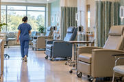 Synthetic editorial illustration. A nurse seen from behind walks through a bright treatment room lined with empty reclining chairs. Plain clear fluid pouches hang from IV stands beside dark pump displays and privacy curtains.