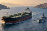 Synthetic editorial illustration. A large dark-hulled tanker and a smaller grey patrol vessel move through a narrow sea passage, leaving gentle wakes. Barren rocky shores frame the water in warm hazy light.