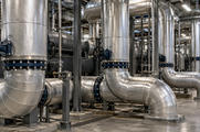 Synthetic editorial illustration. A mechanical plant room. Large silver-insulated pipes bend between floor mounts and overhead runs, with dark handwheel valves, bolted flanges and metal supports repeating into the room.