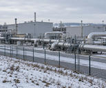 Synthetic editorial illustration. A winter gas compressor station. Large grey buildings and substantial silver pipework stand behind a mesh fence amid snow-dusted ground and leafless trees.