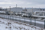 Synthetic editorial illustration. A winter gas compressor station. Large grey buildings and substantial silver pipework stand behind a mesh fence amid snow-dusted ground and leafless trees.