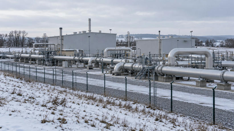 Synthetic editorial illustration. A winter gas compressor station. Large grey buildings and substantial silver pipework stand behind a mesh fence amid snow-dusted ground and leafless trees.