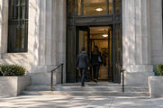 Synthetic editorial illustration. Two rear-view adults enter a formal stone institutional building through a tall doorway in morning light.