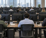 Synthetic editorial illustration. A commodity trading office. Traders seen from behind sit at rows of desks with monitor backs and mounting arms facing the camera, with daylight windows across the far wall.