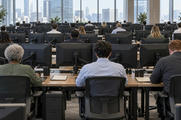 Synthetic editorial illustration. A commodity trading office. Traders seen from behind sit at rows of desks with monitor backs and mounting arms facing the camera, with daylight windows across the far wall.