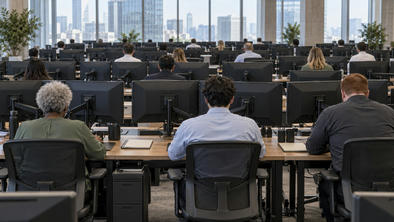 Synthetic editorial illustration. A commodity trading office. Traders seen from behind sit at rows of desks with monitor backs and mounting arms facing the camera, with daylight windows across the far wall.
