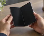Synthetic editorial illustration. Two hands hold a partially unfolded phone above a wooden desk. Its two dark display halves meet at a visible central hinge, with a plain mug, plant and notebook around the edges.