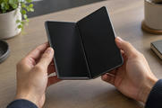 Synthetic editorial illustration. Two hands hold a partially unfolded phone above a wooden desk. Its two dark display halves meet at a visible central hinge, with a plain mug, plant and notebook around the edges.