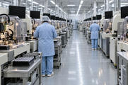 Synthetic editorial illustration. A chip packaging hall. Rows of guarded precision machines and component trays line a long aisle, with two operators in pale blue smocks seen from behind and dark monitors above the machines.