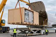 Synthetic editorial illustration. A crane suspends a large plain wooden crate containing wrapped machinery beside an open cargo-aircraft hold. Two small rear-facing workers guide the load above a lifting platform under overcast daylight.