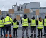 Synthetic editorial illustration. Workers in plain fluorescent vests stand with their backs to the viewer outside a metal factory gate. Several hold blank cardboard placards beneath an overcast sky, with industrial pipes and sheds beyond.