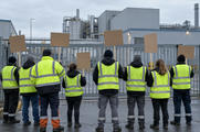 Synthetic editorial illustration. Workers in plain fluorescent vests stand with their backs to the viewer outside a metal factory gate. Several hold blank cardboard placards beneath an overcast sky, with industrial pipes and sheds beyond.