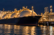 Synthetic editorial illustration. An LNG terminal. A large dark-hulled carrier with spherical tanks lies beside illuminated loading arms and pipework, with warm industrial lights reflecting across the water under a deep blue night sky.