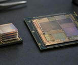 Synthetic editorial illustration. A small stack of thin reflective silicon layers sits on a dark package beside a larger patterned processor die. Both rest on a textured black anti-static mat under soft inspection light.