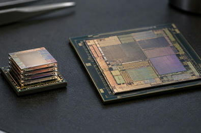 Synthetic editorial illustration. A small stack of thin reflective silicon layers sits on a dark package beside a larger patterned processor die. Both rest on a textured black anti-static mat under soft inspection light.