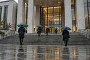 Synthetic editorial illustration. A modern courthouse entrance. People seen from behind climb broad wet stone steps beneath plain umbrellas toward a lit glass entrance as rain falls.
