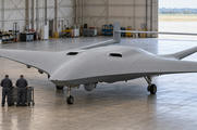 Synthetic editorial illustration. An unmarked broad blended-wing aircraft with twin tail booms, a straight joining tailplane and upright fins stands in a daylit hangar. Two small workers seen from behind stand beside a tool trolley.