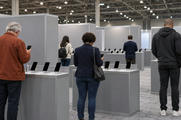 Synthetic editorial illustration. Visitors of varied ages and builds viewed from behind test dark-screen phones at plain grey counters. Modular grey booths, carpeted aisles and exposed roof trusses define the technology exhibition hall.